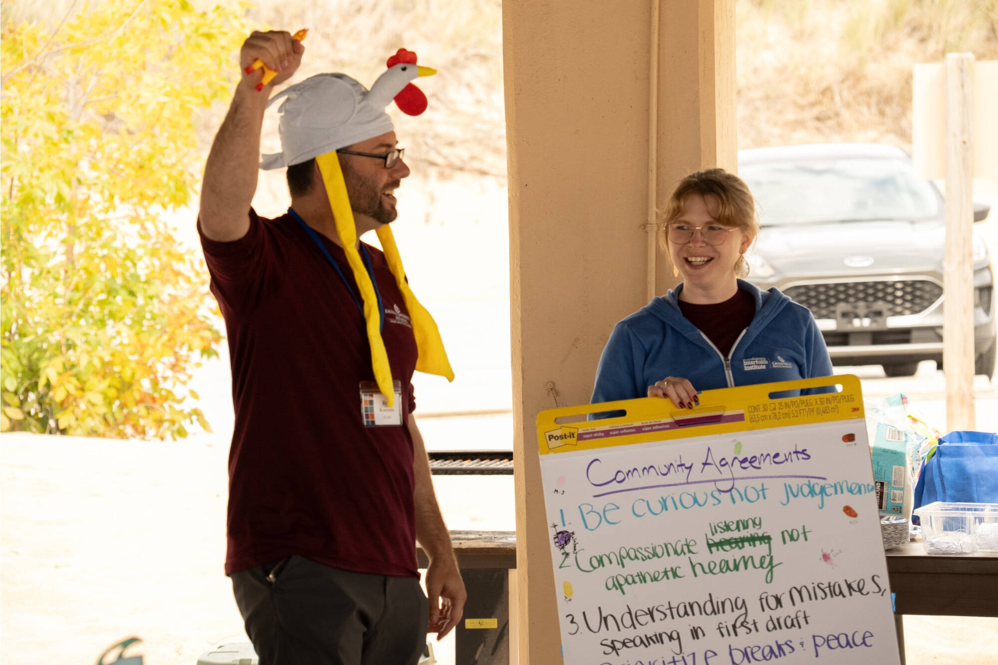 A man in a chicken hat squeezing a rubber chicken with a woman holding a large poster of community agreements in the background.